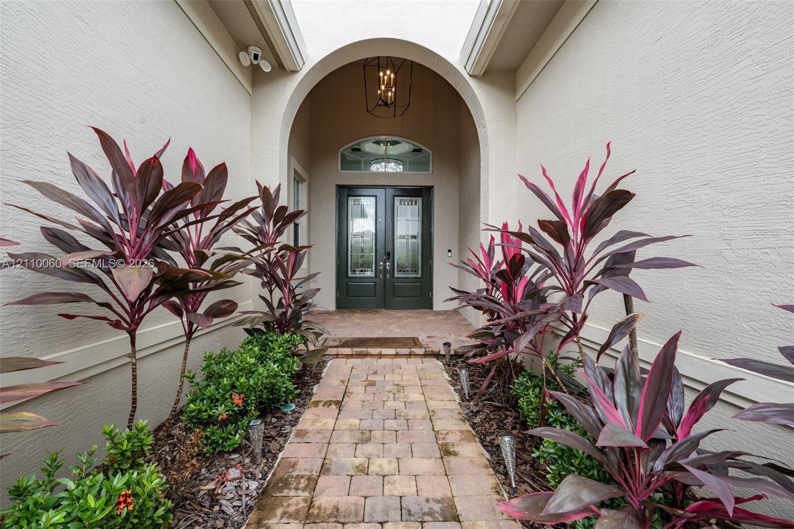 Arched entryway featuring a paver walkway, double-door entry with arched transom window, built-in geometric lantern-style lighting, and mature landscape plantings