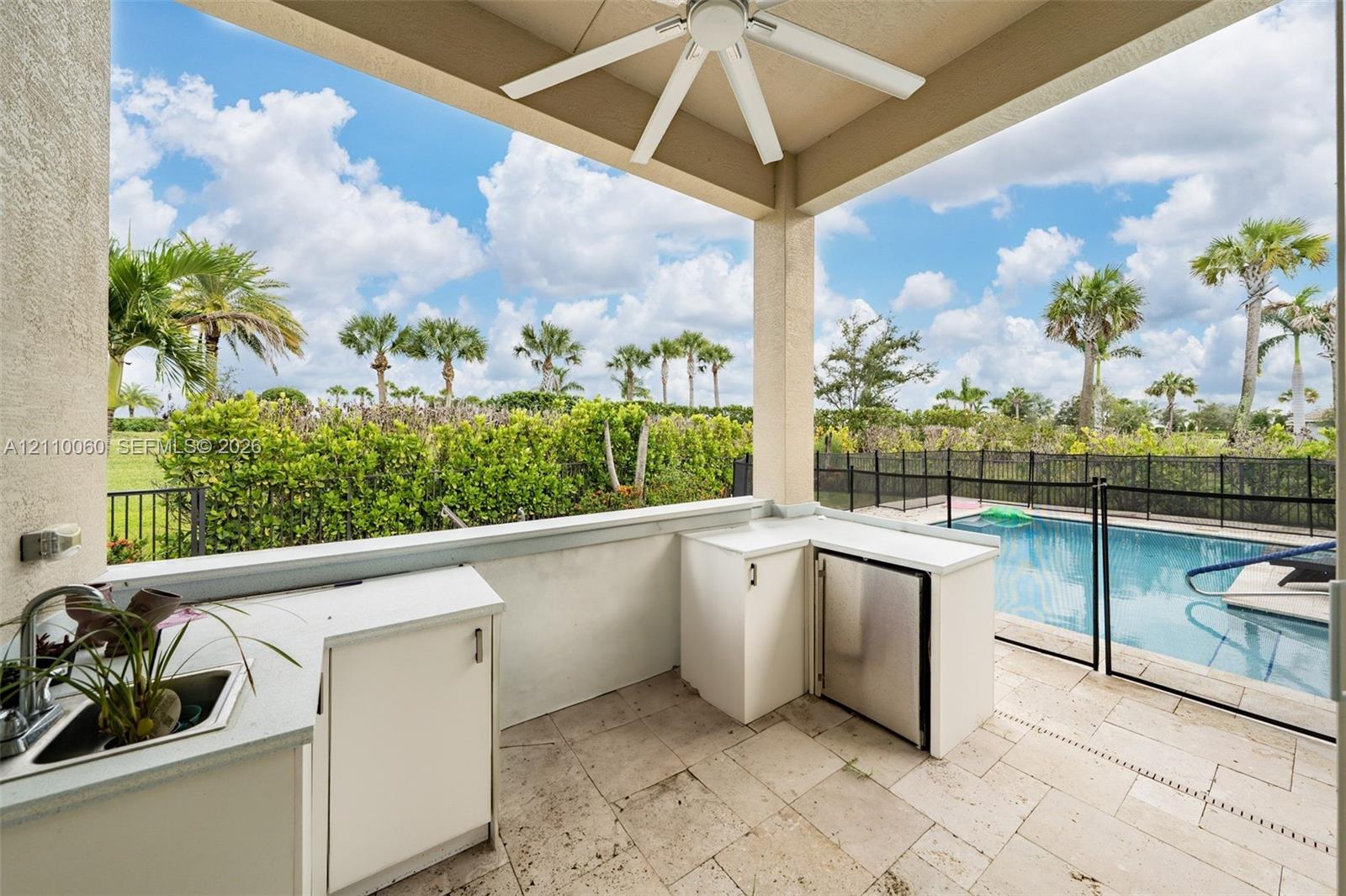 Covered outdoor kitchen featuring light-toned cabinetry, solid surface countertops, stainless steel refrigeration, and a sink, complete with stone-tiled flooring and a ceiling fan, overlooking a fenced pool