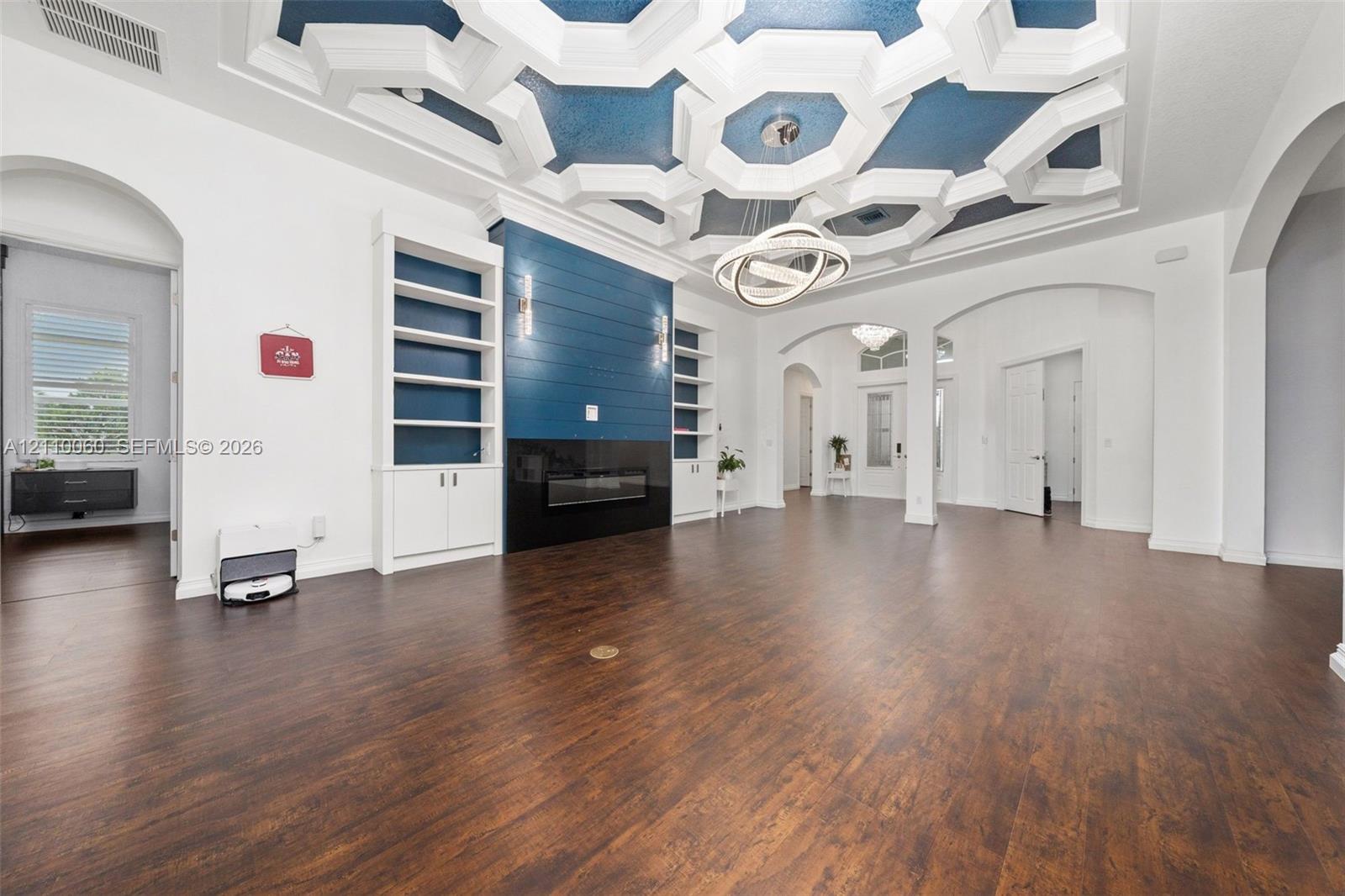 Geometric coffered ceiling with inset blue accents, wood-finish flooring, built-in floor-to-ceiling cabinetry, shiplap fireplace surround, and modern circular tiered chandelier