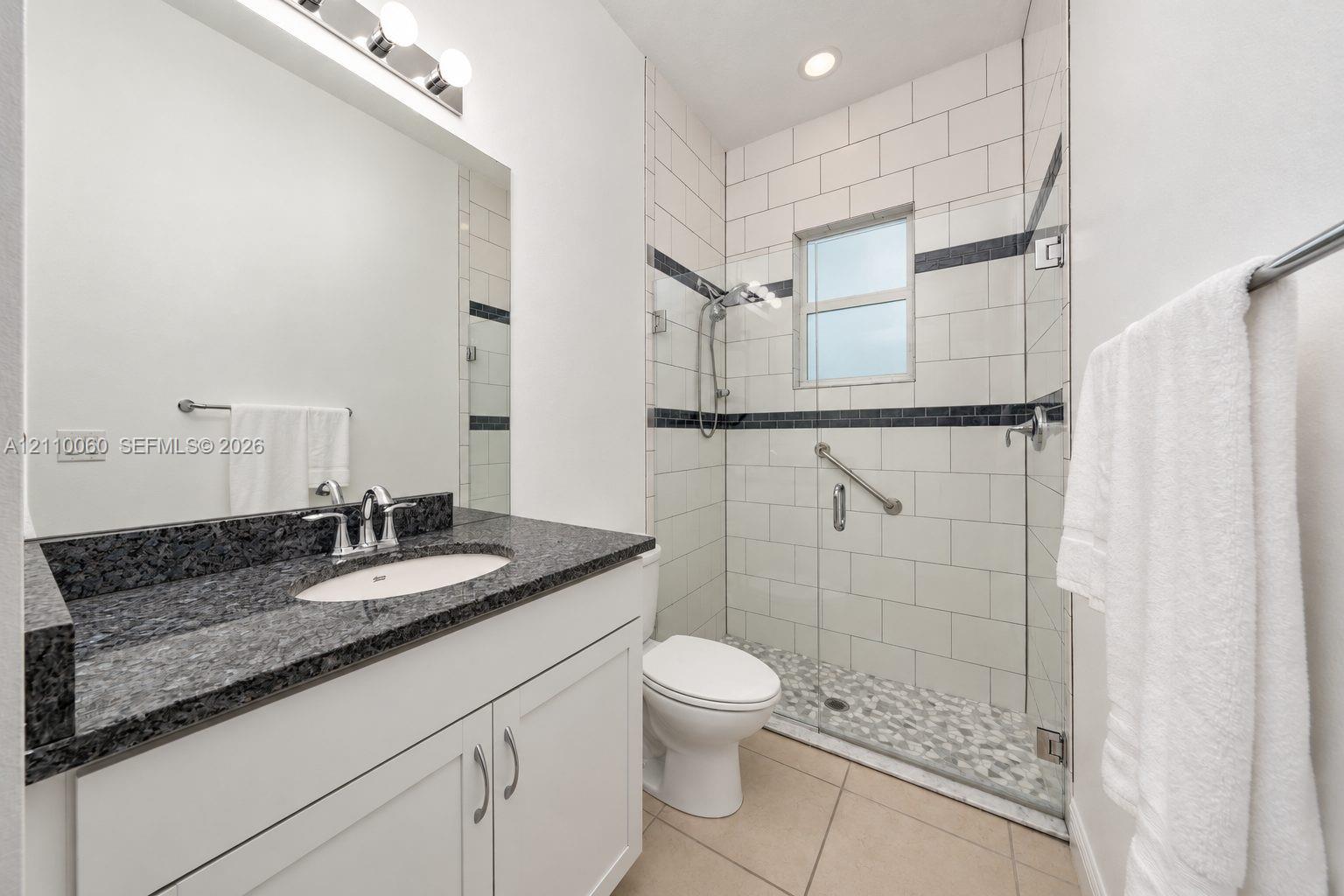 Bathroom featuring a white vanity cabinet with a dark stone countertop, an integrated sink with a chrome faucet, a walk-in shower with a glass enclosure and multi-tone tile accents, and large-format tile flooring
