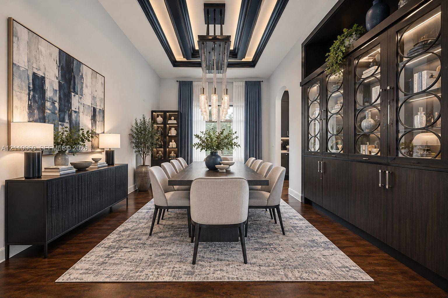 Virtually staged. Formal dining area featuring a tray ceiling with recessed lighting, custom floor-to-ceiling built-in cabinetry with glass-paneled display doors, dark wood-finish flooring, and a multi-tiered crystal pendant light fixture
