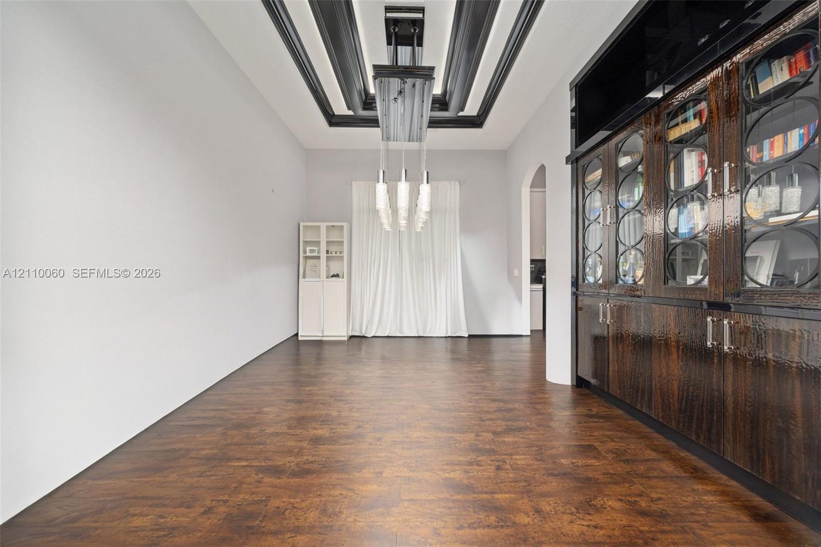 Wood-finish flooring, dramatic coffered ceiling with black detailing, built-in floor-to-ceiling cabinetry with glass-panel doors, and an elongated multi-light pendant fixture