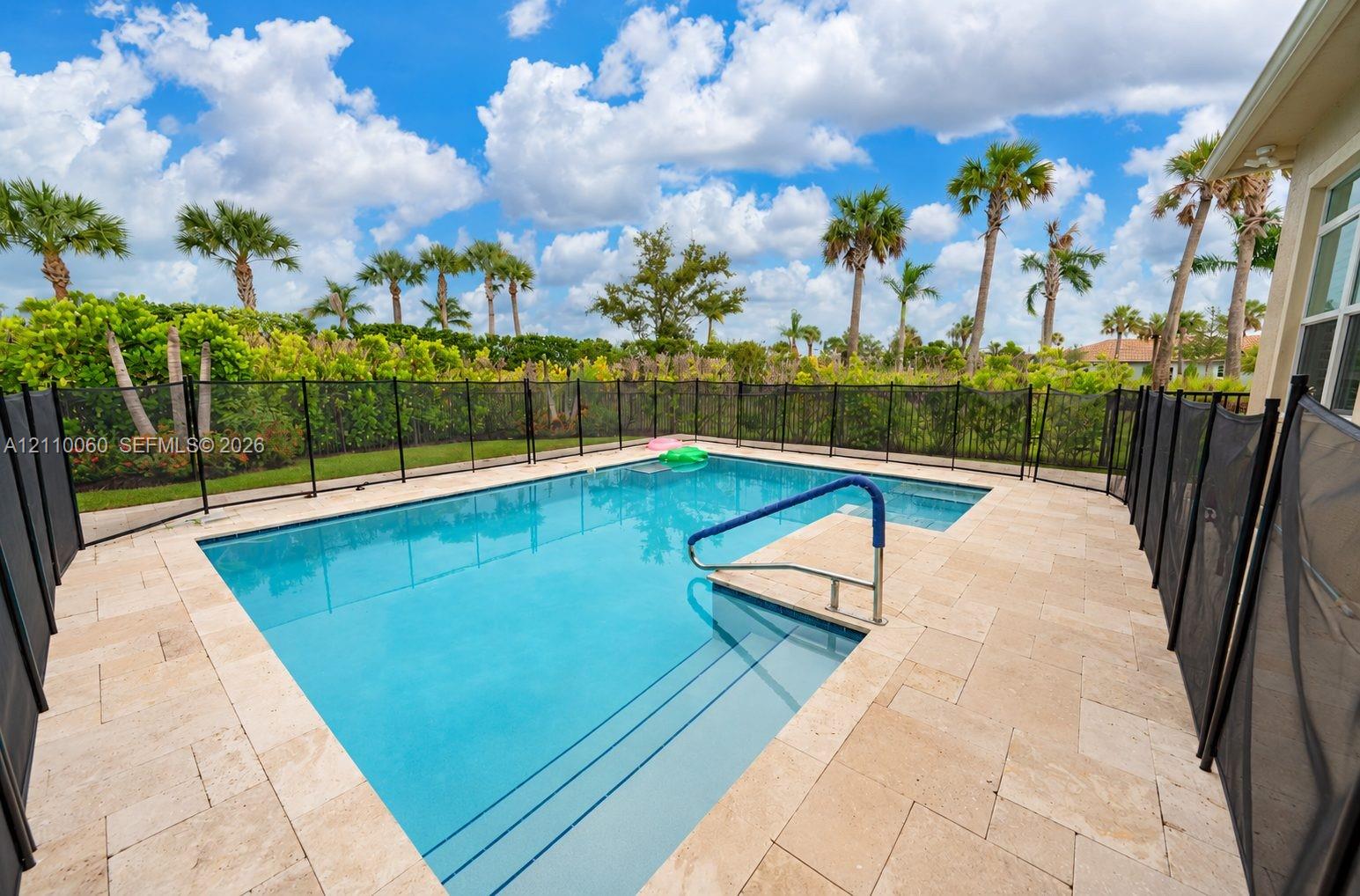 In-ground pool with built-in stairs, tan stone paver decking, black mesh safety fencing, and mature palm tree landscaping