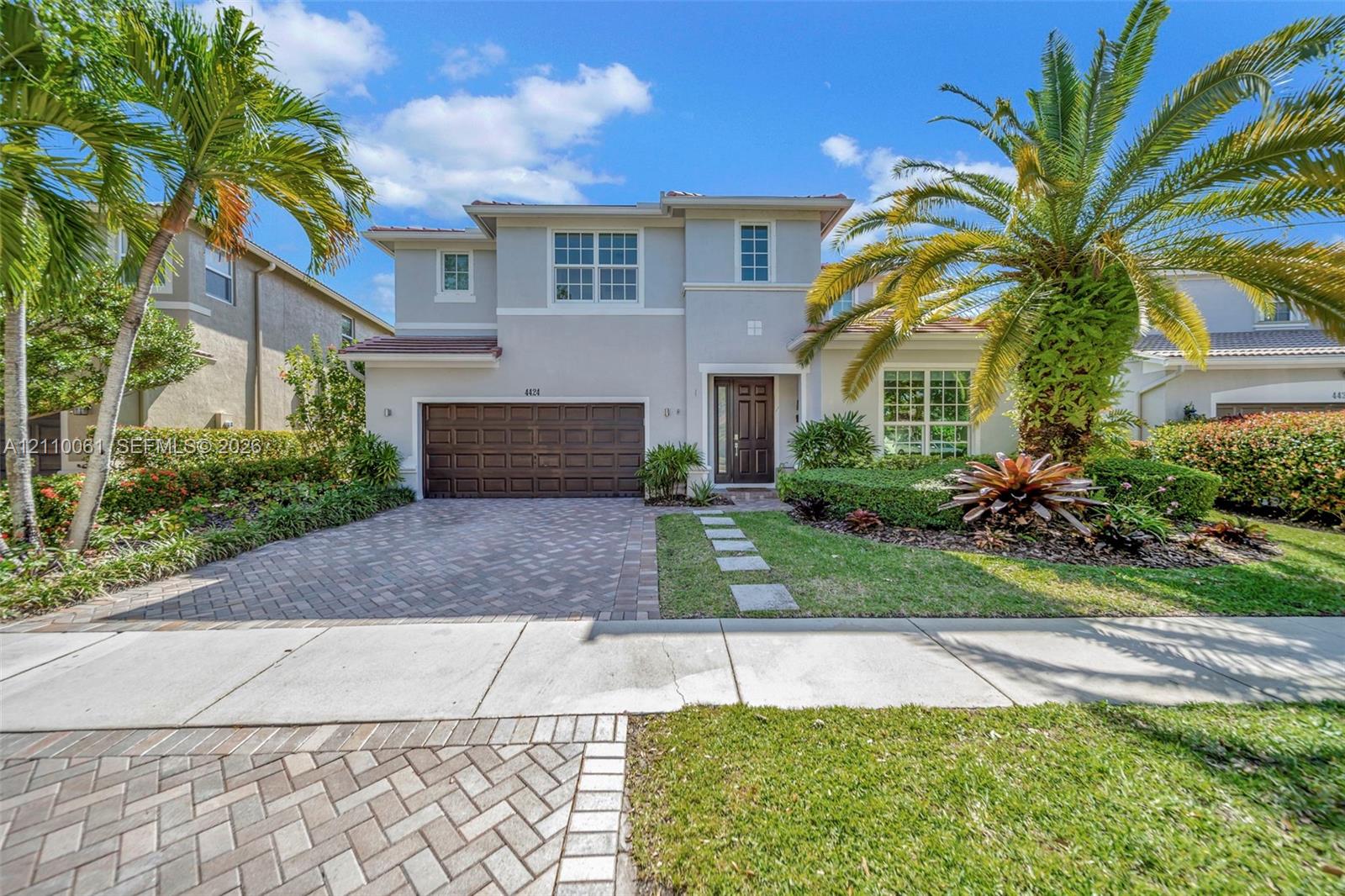 Two-story residence featuring a stucco facade, paved driveway, multi-panel garage door, and a manicured front lawn with mature palm trees Two-story residence featuring a stucco facade, paved driveway, multi-panel garage door, and a manicured front lawn with mature palm trees