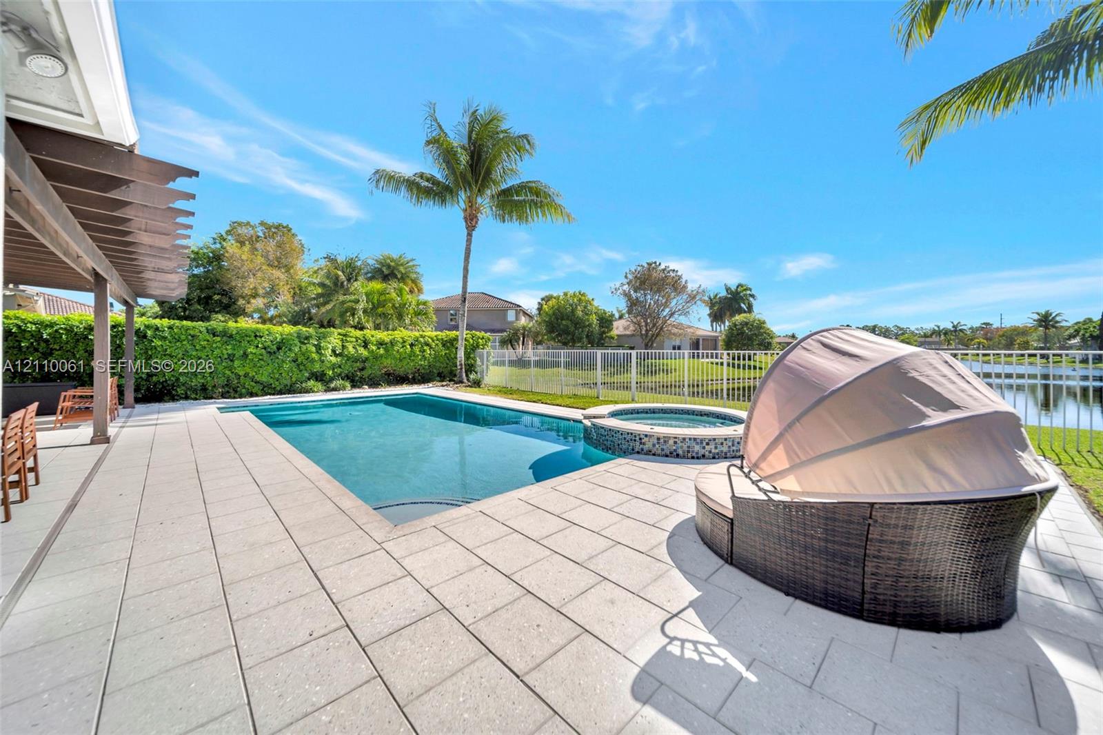 Swimming pool and attached spillover spa with mosaic tile accents, extensive concrete paver pool deck, covered pergola, and perimeter metal fencing