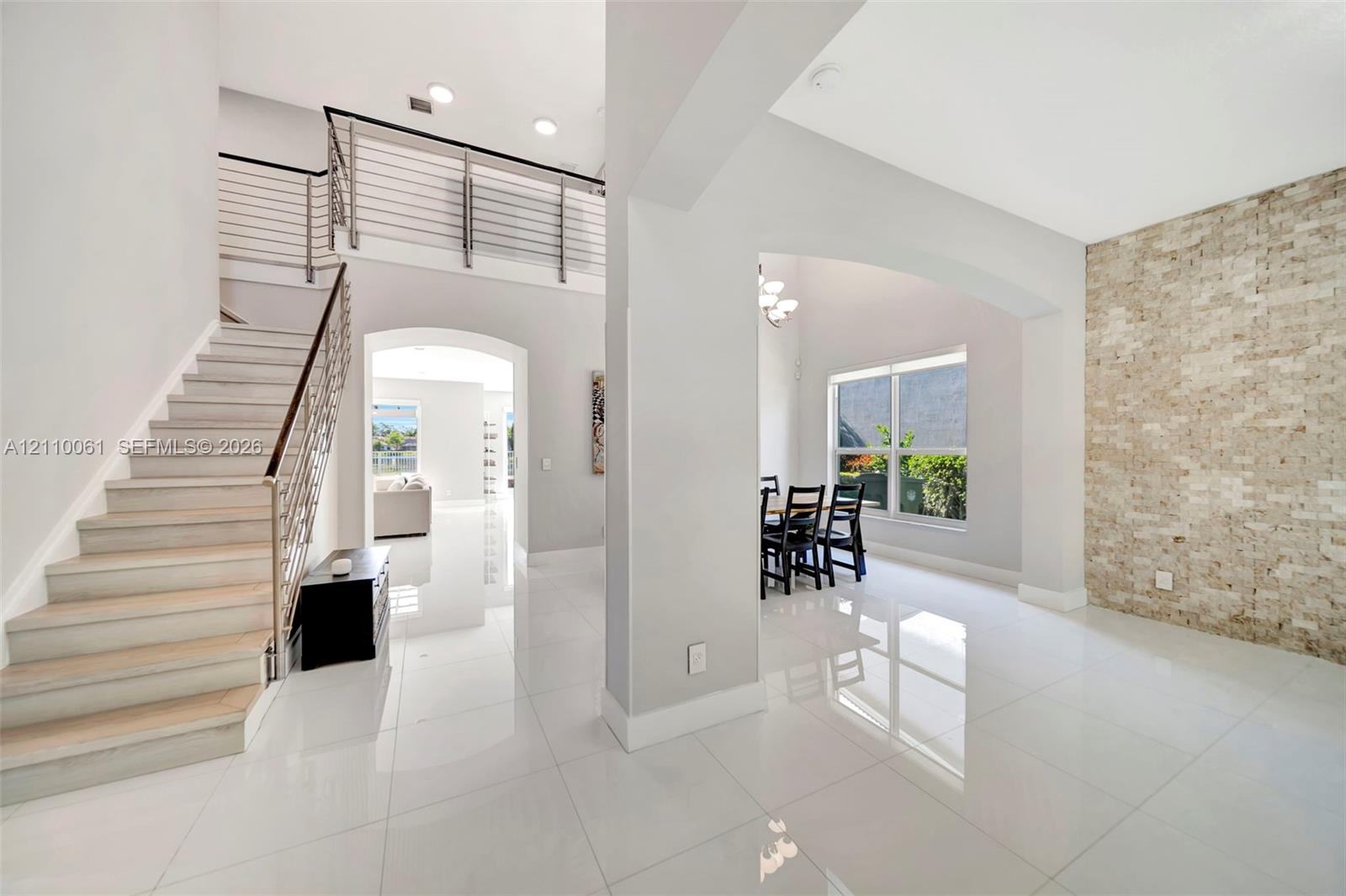 Open-concept interior featuring large-format tile flooring, a floor-to-ceiling textured stone accent wall, a staircase with custom metal railing, and high ceilings with recessed lighting