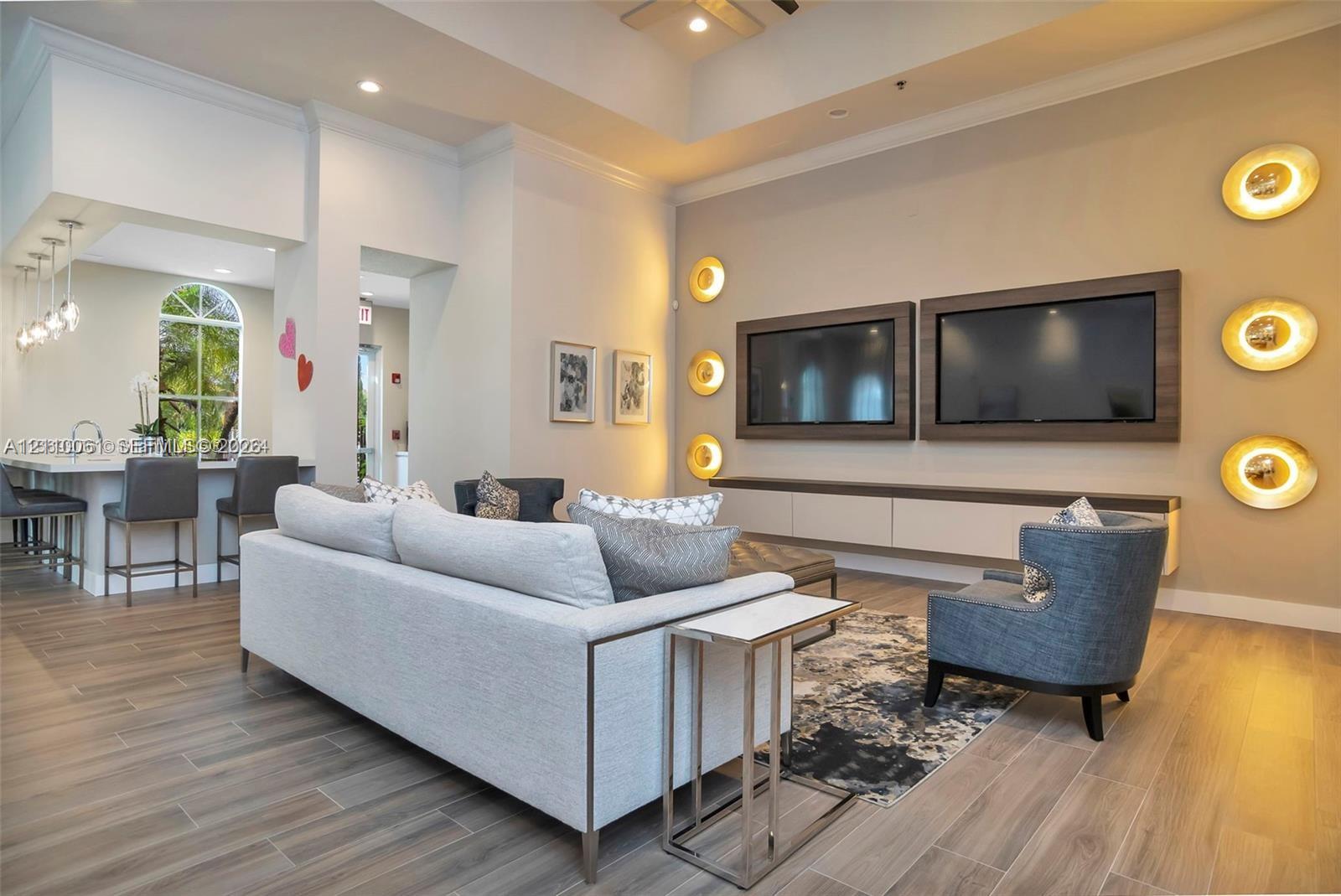 Open-concept living space featuring high tray ceilings, wood-finish flooring, a wall-mounted media console with built-in cabinetry, custom circular wall sconces, and a breakfast bar with pendant lighting