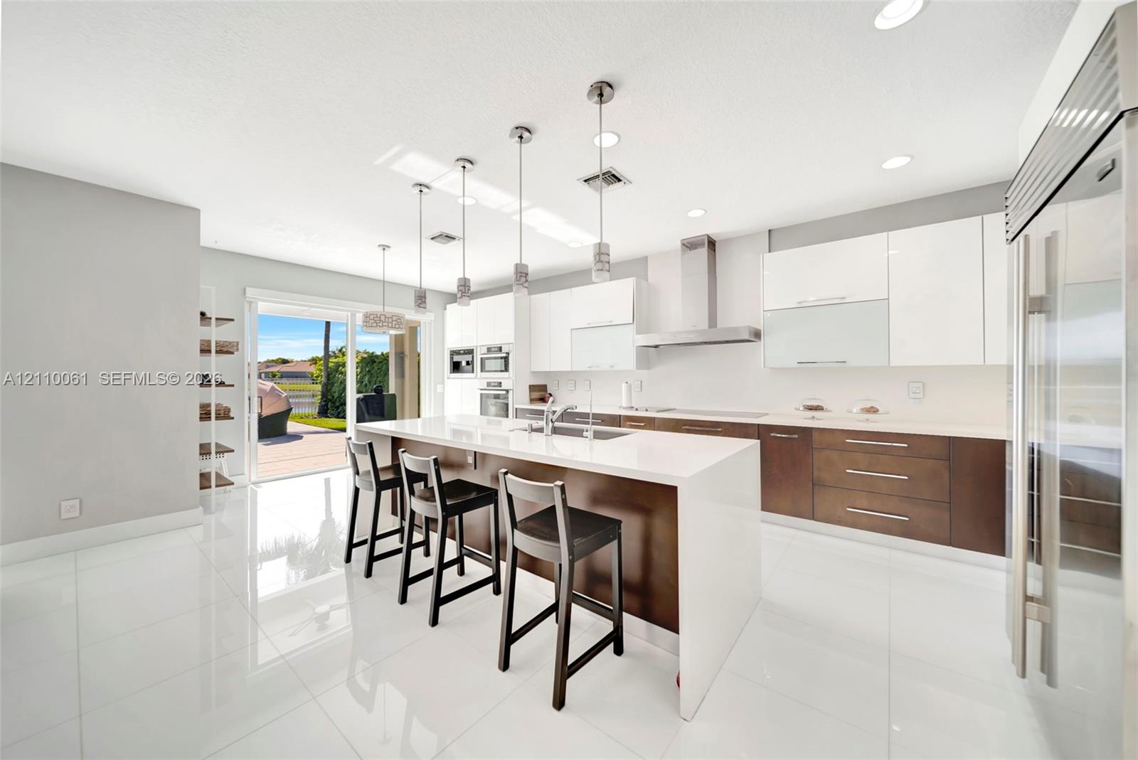 Contemporary kitchen featuring an expansive waterfall-edge island, two-tone cabinetry with gloss-finish uppers, stainless steel professional-grade appliances, recessed lighting, and large-format tile flooring