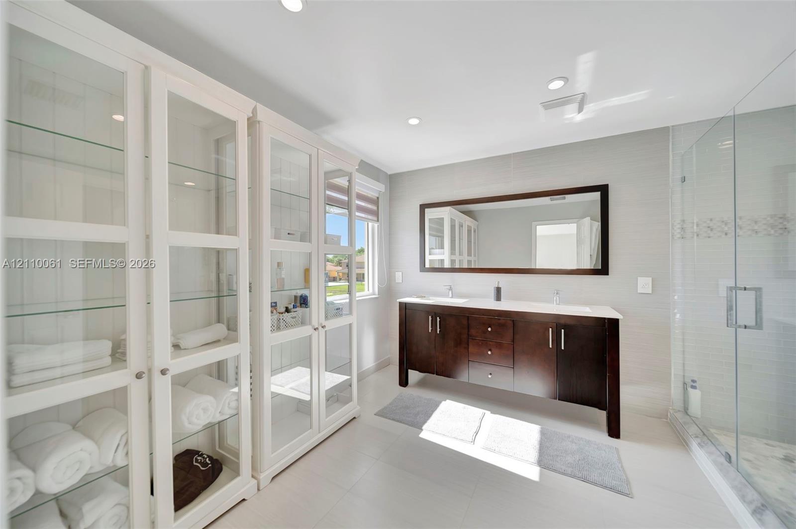 Double vanity with a dark wood-finish cabinet and surface countertop, glass-enclosed shower with tile surround, and extensive built-in cabinetry featuring glass-front doors