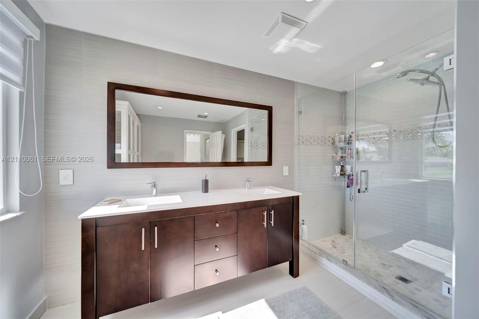 Double vanity with rectangular undermount sinks and light-toned solid surface countertop, wood-finish cabinetry with metallic hardware, and walk-in glass-enclosed shower featuring a handheld fixture and tiled surround