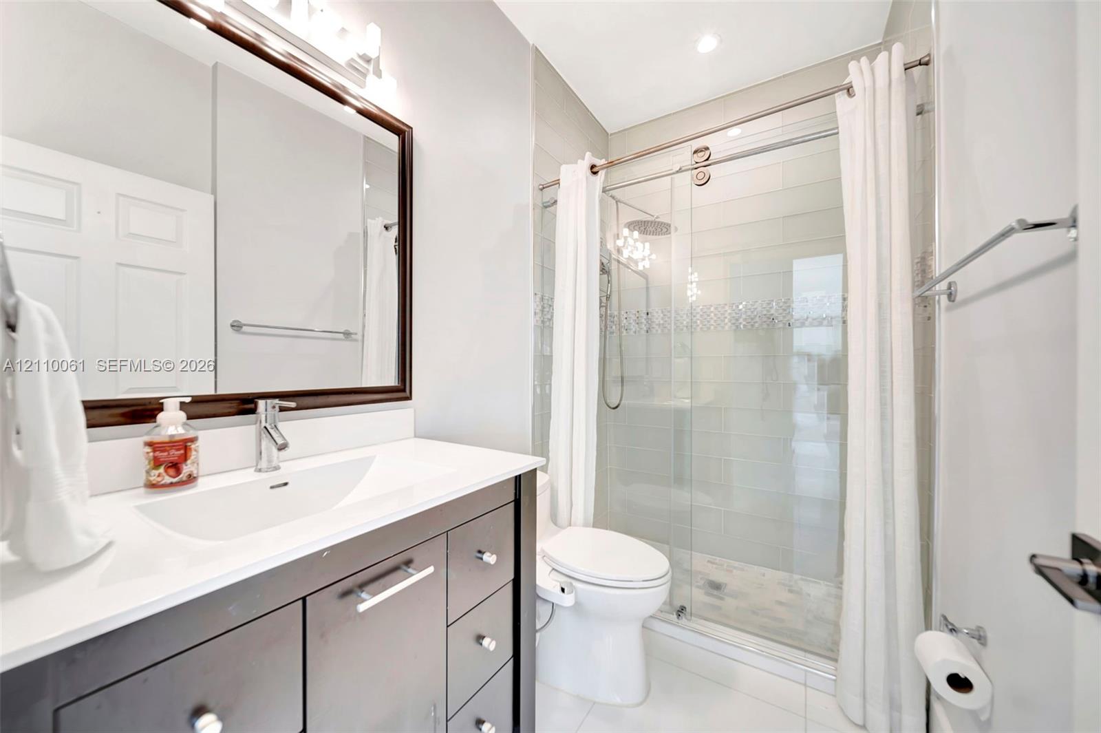 Bathroom featuring a dark-finish vanity with a rectangular sink, quartz countertop, framed wall mirror, and a walk-in shower with sliding glass doors and tiled surround