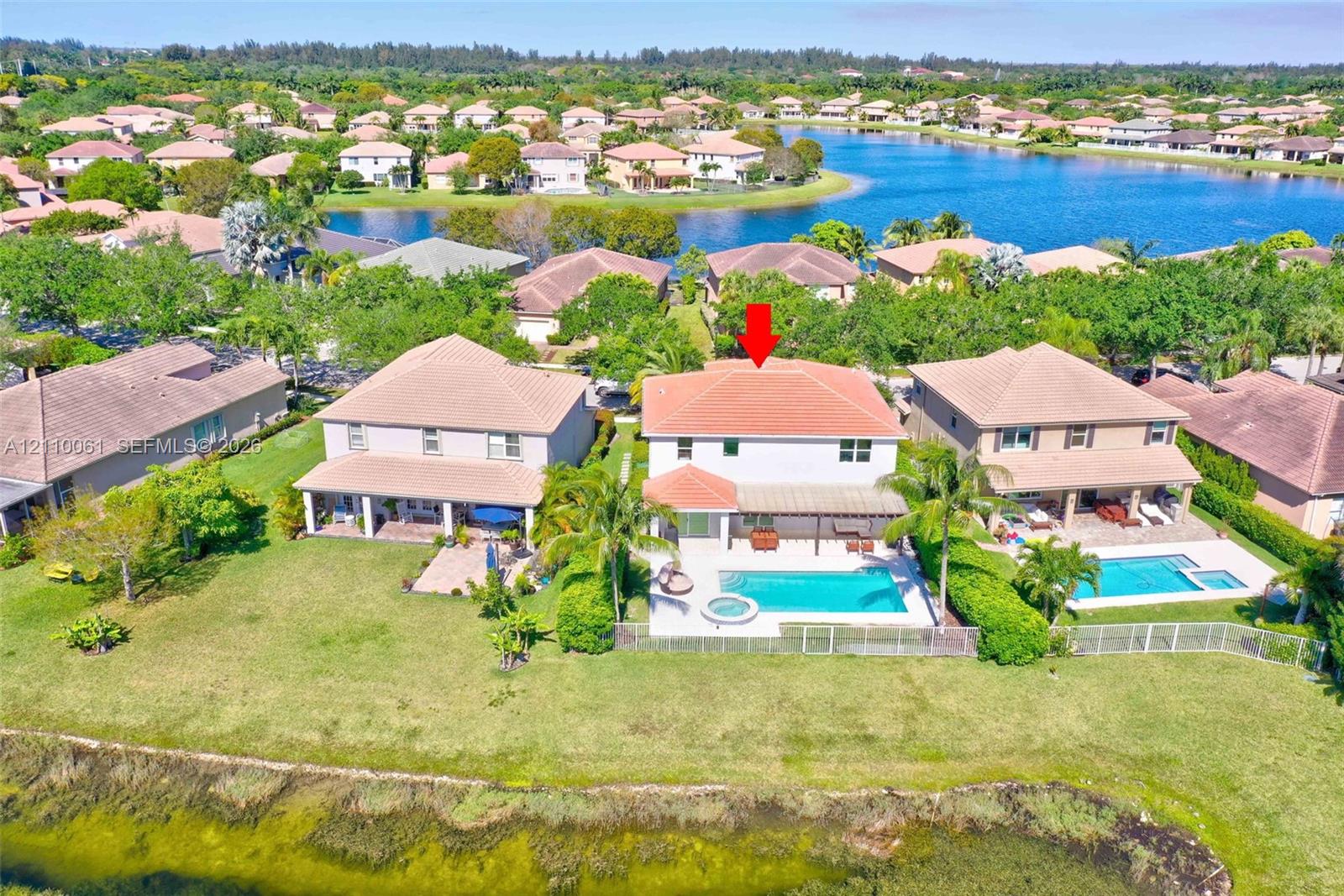 Waterfront residence featuring a two-story architectural design with a terracotta tile roof, expansive manicured lawn, mature landscaping, in-ground swimming pool with surrounding hardscaping, and direct waterfront access overlooking the lake community