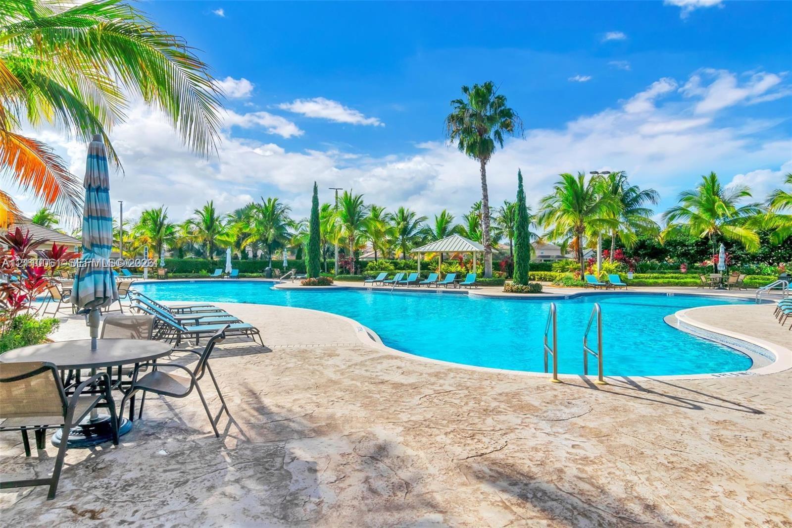 Resort-style swimming pool featuring a sprawling stone-paved deck, integrated metal ladders, shaded pavilion, and lush palm tree landscaping
