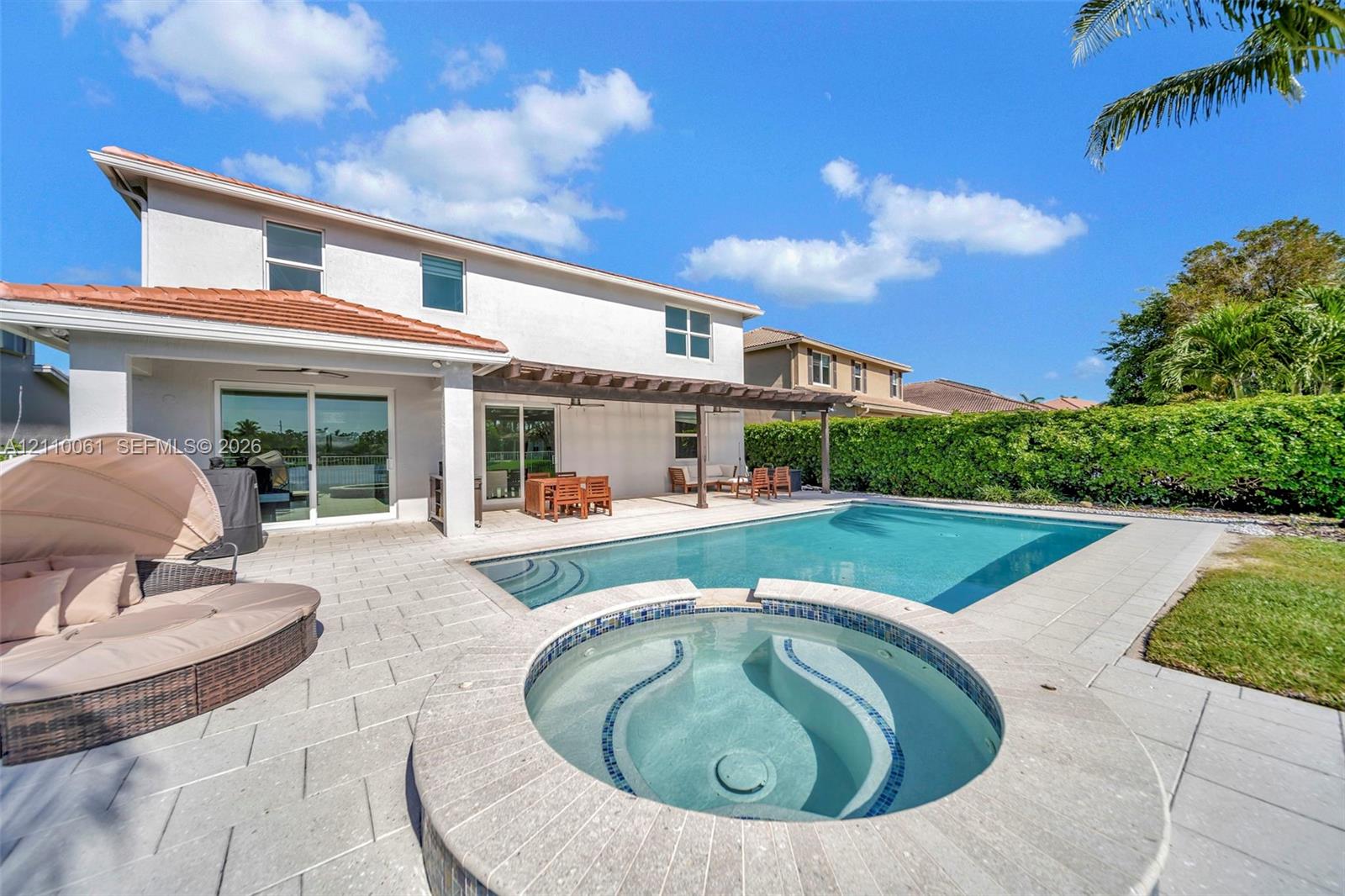 Rear exterior featuring a covered patio with pergola, a rectangular swimming pool, a raised integrated spa, and light-toned stone-finish paver decking