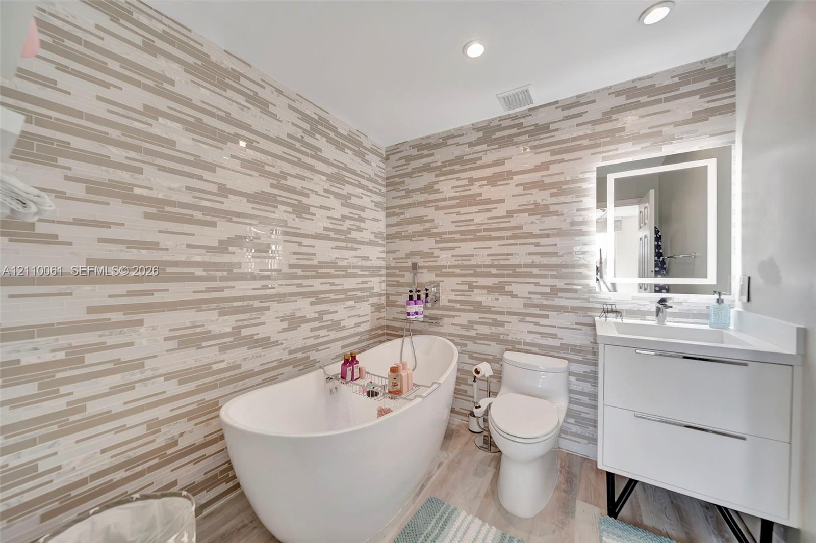 Bathroom featuring floor-to-ceiling multi-tonal linear tile walls, a freestanding soaking tub with handheld sprayer, a vanity with a white countertop and metal legs, and an integrated LED-backlit mirror