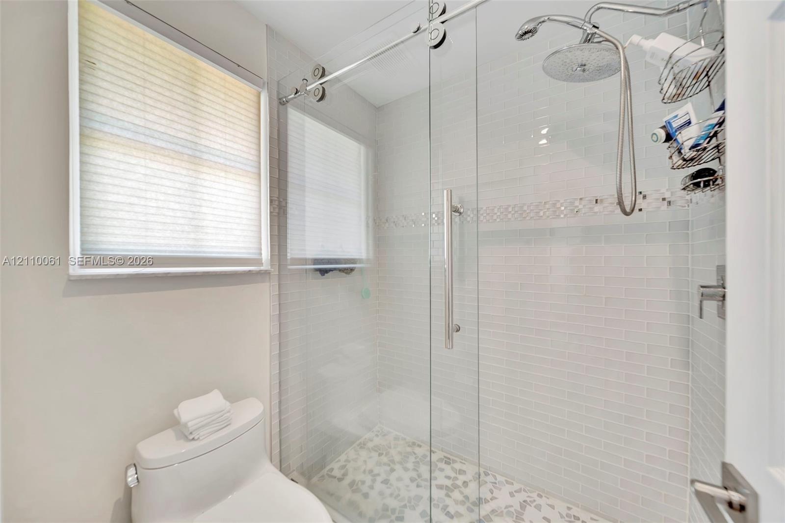 Walk-in shower featuring a glass sliding door, polished metal rain showerhead with handheld attachment, horizontal white tile walls with a decorative mosaic inlay, and textured stone floor tiling