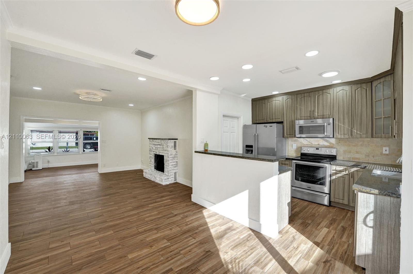 Kitchen featuring wood-finish cabinetry, stone-texture backsplash, stainless steel appliances, and a breakfast bar, complemented by wood-finish flooring and a whitewashed brick fireplace Kitchen featuring wood-finish cabinetry, stone-texture backsplash, stainless steel appliances, and a breakfast bar, complemented by wood-finish flooring and a whitewashed brick fireplace