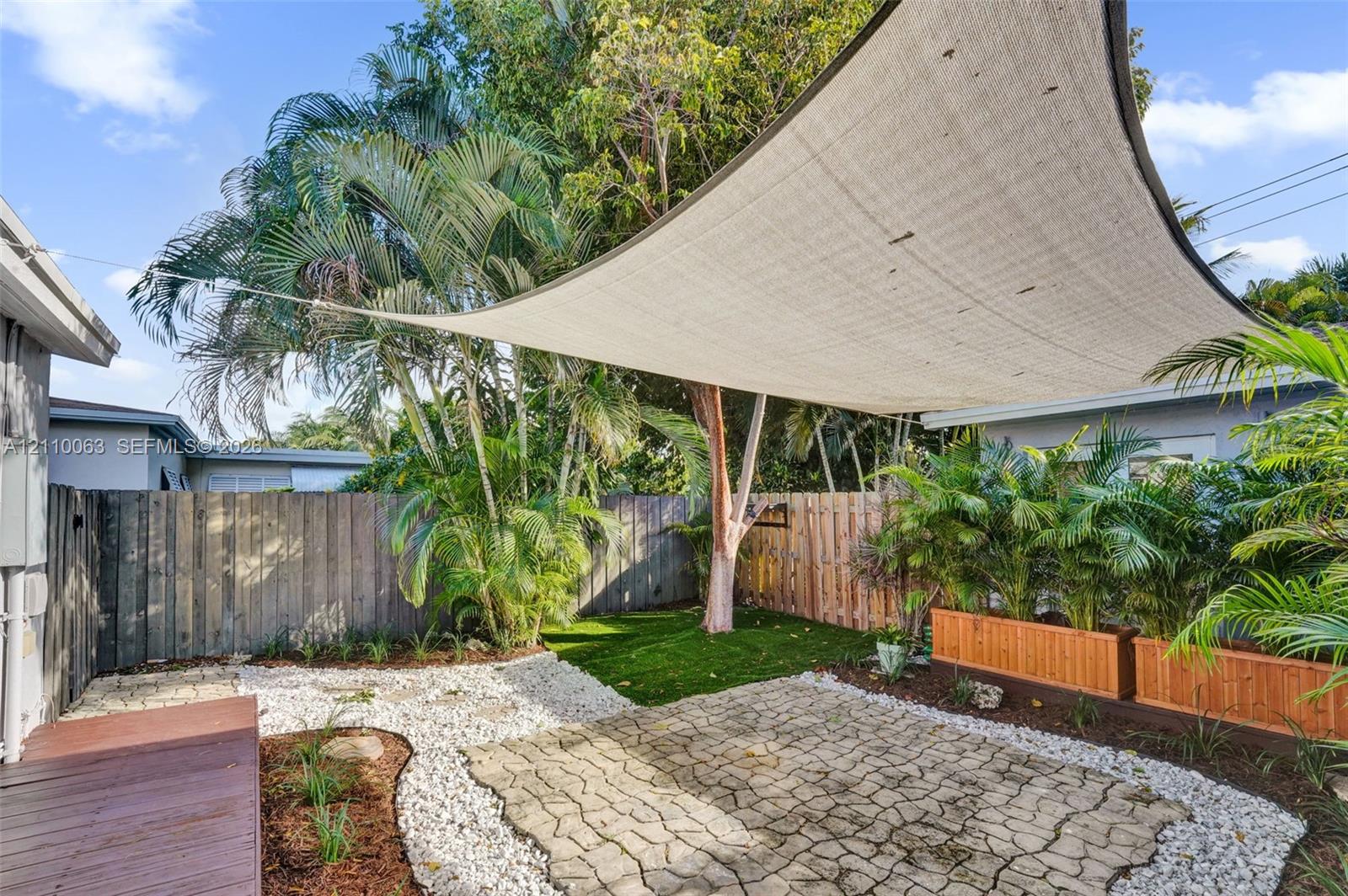 Outdoor patio area featuring a stone paver floor, artificial turf, perimeter wood fencing, a fabric shade sail, and integrated raised wooden planters