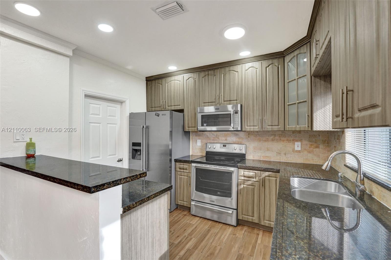 Kitchen featuring wood-finish cabinetry, dark stone countertops, stainless steel appliances, a tiled backsplash, and wood-finish flooring