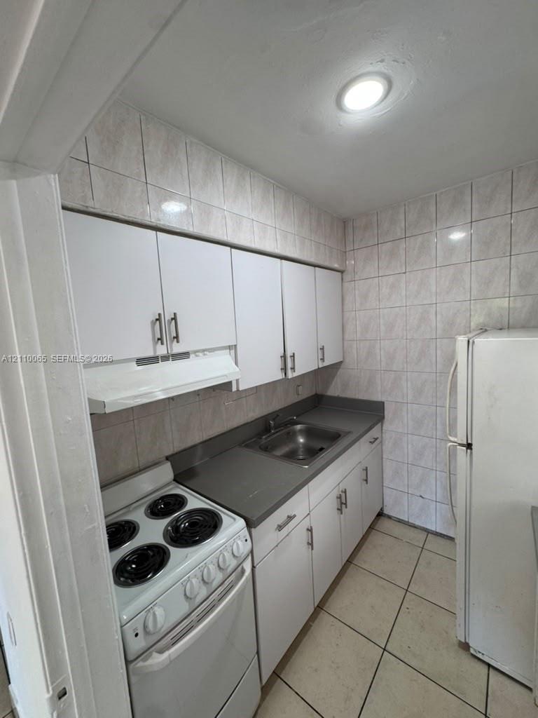 Kitchen featuring white cabinetry with metallic hardware, solid-surface countertops, a stainless steel basin sink, ceramic wall tiling, and ceramic floor tiling
