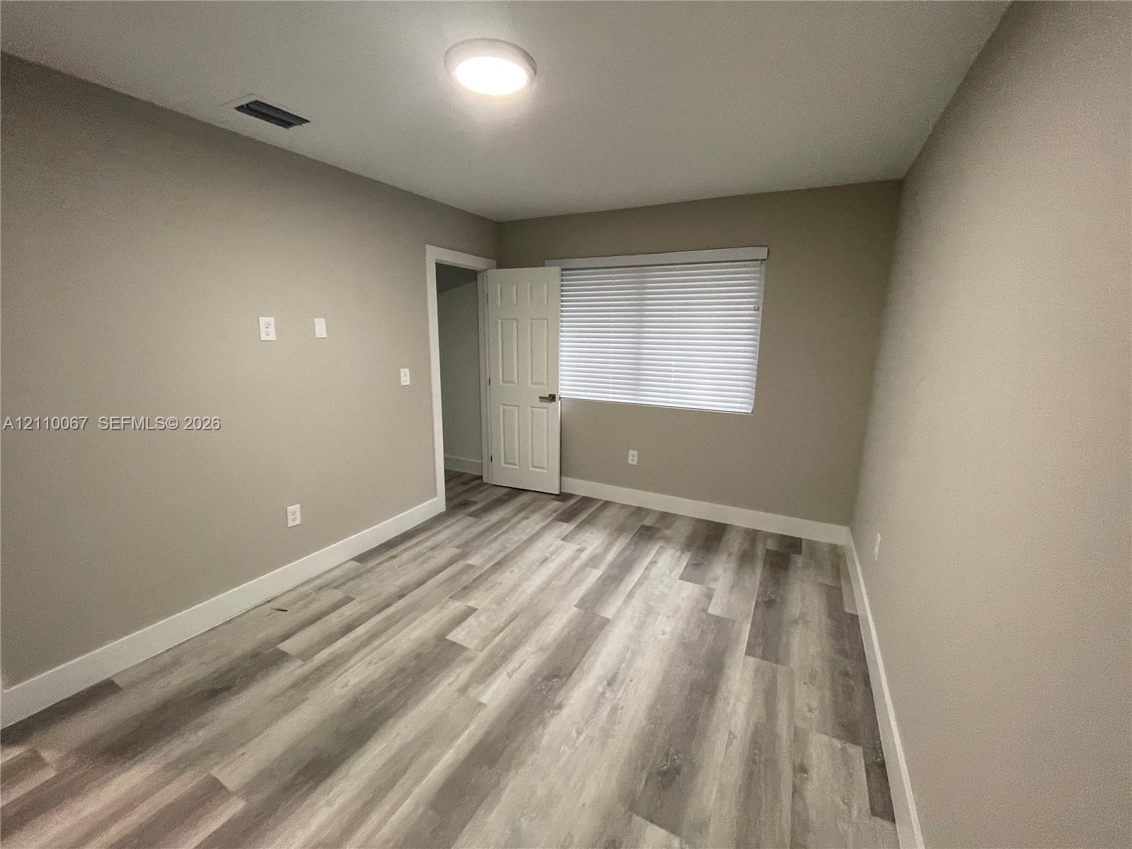 Neutral-toned interior space featuring wood-finish flooring, a six-panel door, and a horizontal window with adjustable blinds