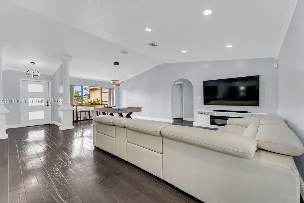 Open-concept interior featuring vaulted ceilings, dark wood-finish flooring, recessed lighting, architectural columns, and a built-in media console with an integrated fireplace