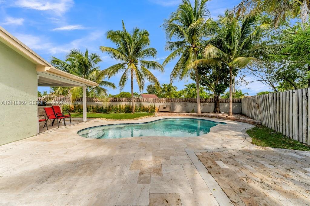 Outdoor living area featuring a custom-shaped swimming pool, extensive stone paver patio, covered porch, and mature palm trees enclosed by wooden fencing