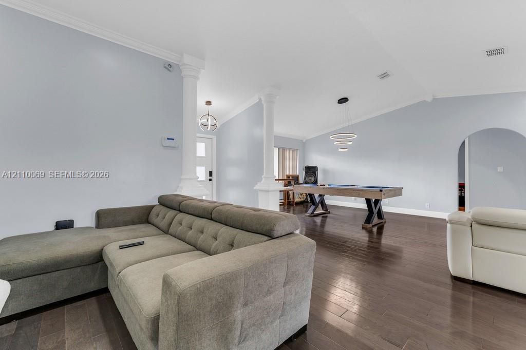 Open-concept interior featuring vaulted ceilings, dark wood-finish flooring, classic architectural columns, multi-tiered ring lighting, and a signature arched entry