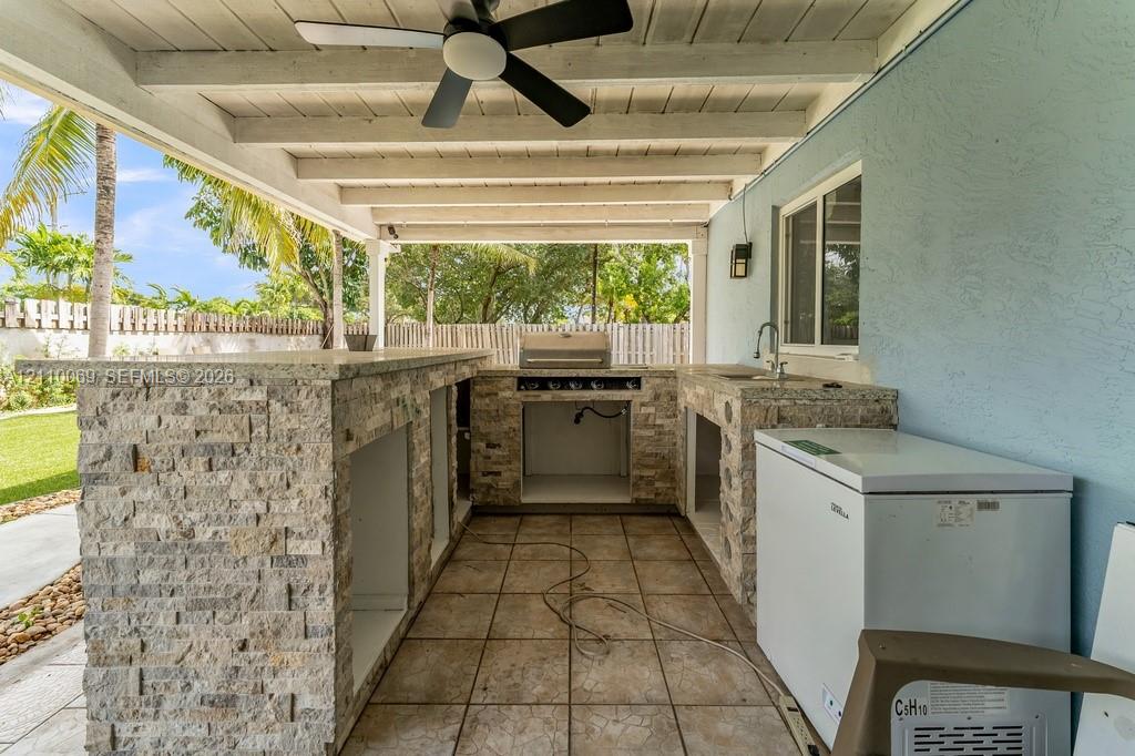 Covered outdoor kitchen featuring stone veneer cabinetry, solid surface countertop, built-in grill, integrated sink, and overhead ceiling fan