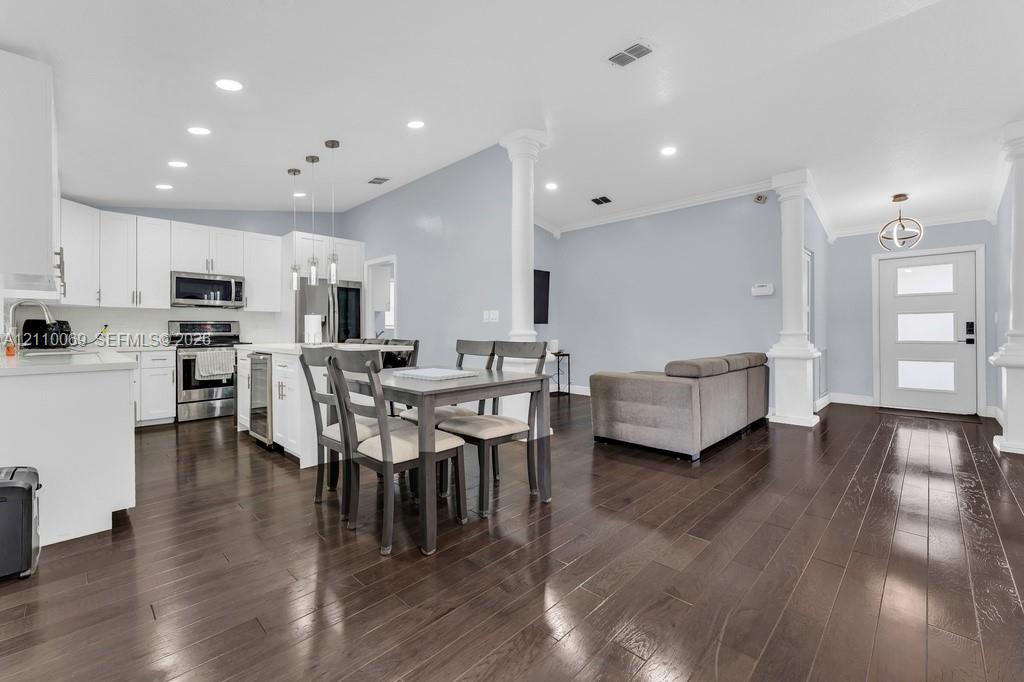 Open-concept kitchen and living area featuring dark wood-finish flooring, white cabinetry, stainless steel appliances, decorative structural columns, and recessed lighting