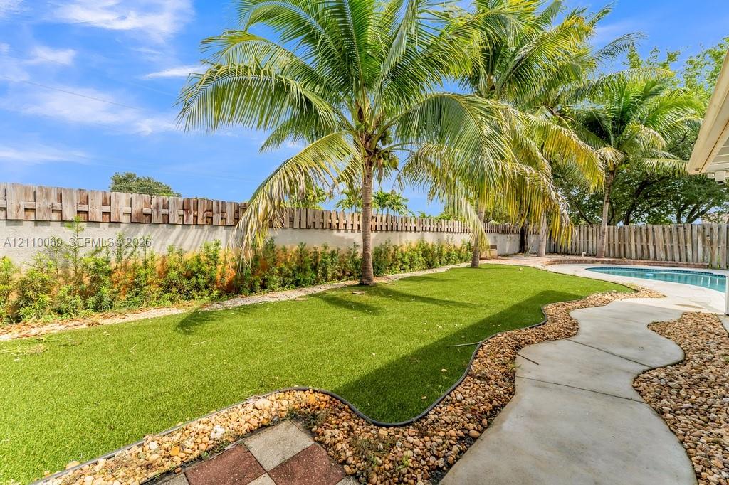 Fenced backyard featuring synthetic turf, palm trees, stone-pebble landscaping, concrete walkway, and a swimming pool