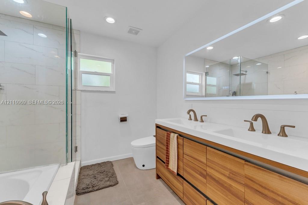 Dual-sink vanity with wood-finish cabinetry, white countertop, brass-tone hardware, large wall-mounted mirror, and glass-enclosed shower with light-toned wall tile
