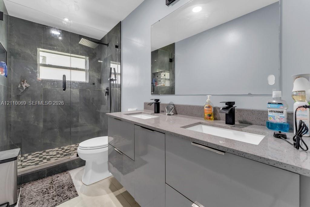 Floating dual-sink vanity with matte black fixtures, engineered stone countertop, full-width mirror, and glass-enclosed shower with floor-to-ceiling tile and rainfall showerhead