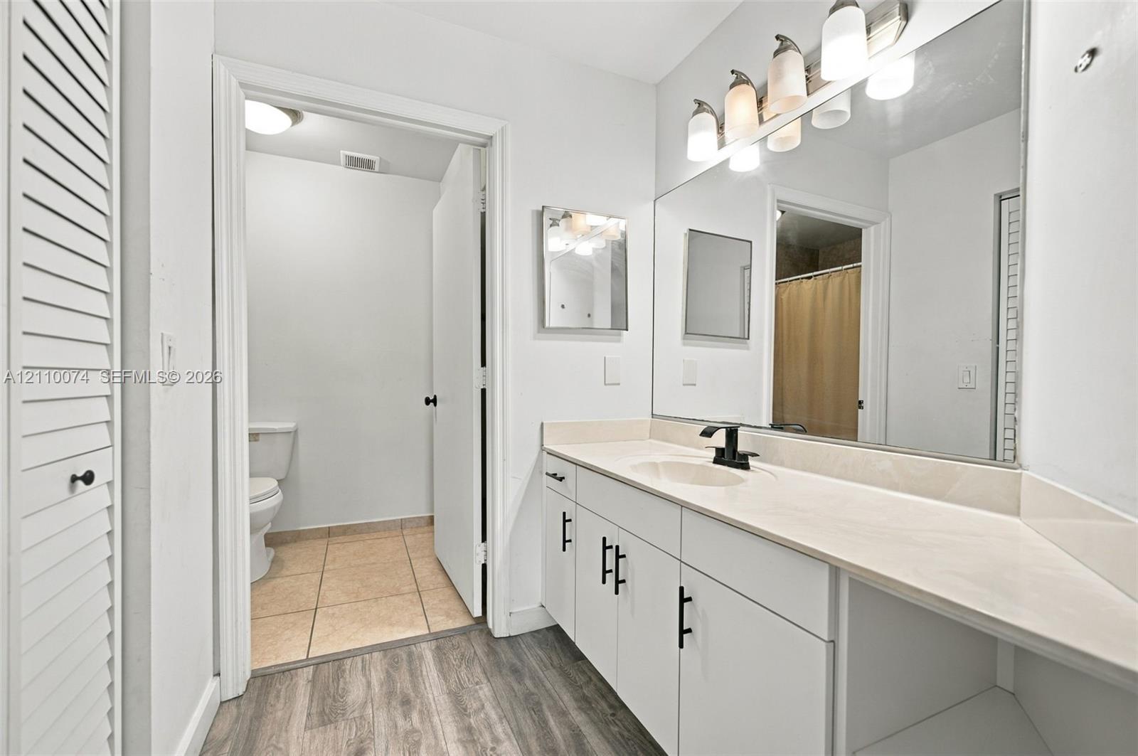 Bathroom vanity featuring a light-toned solid surface countertop, matte black faucet, and white shaker-style cabinetry with matte black hardware, paired with a full-width vanity mirror and wood-finish flooring