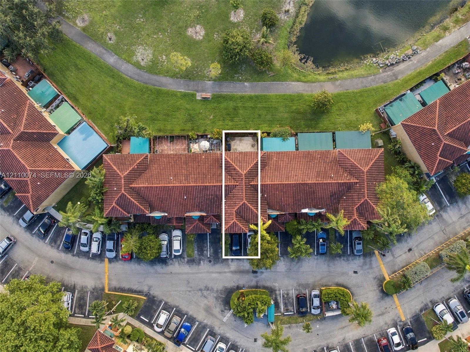 Aerial perspective of multi-family residence featuring a clay tile roof, dedicated parking spaces, and a rear patio area bordering a community pond and walking path