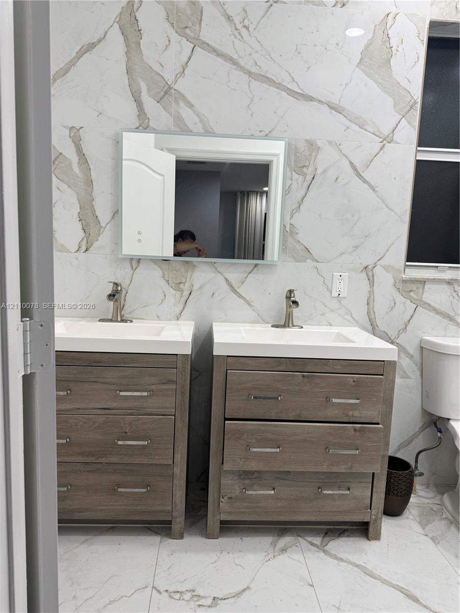 Dual wood-finish vanities with rectangular white sinks, polished metal fixtures, wall-mounted mirror, and floor-to-ceiling large-format tile wall cladding