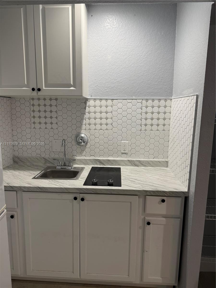 Kitchenette area featuring white shaker-style cabinetry, stone-patterned countertop, drop-in stainless steel sink with dual-handle faucet, integrated electric cooktop, and honeycomb-patterned tile backsplash