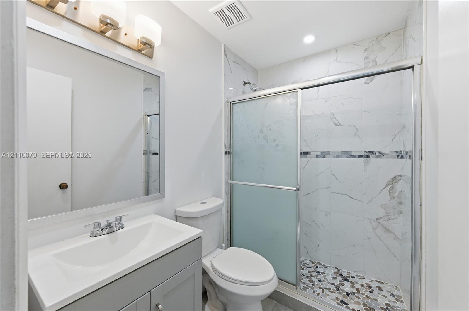 Bathroom featuring a gray vanity with a white countertop, framed wall mirror, three-light fixture, and a glass-enclosed shower with tiled walls and pebble-stone flooring