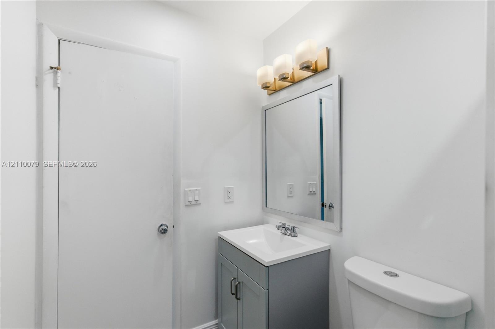 Gray vanity with a white countertop and basin, three-light fixture with glass shades, and a rectangular mirror with a metal frame