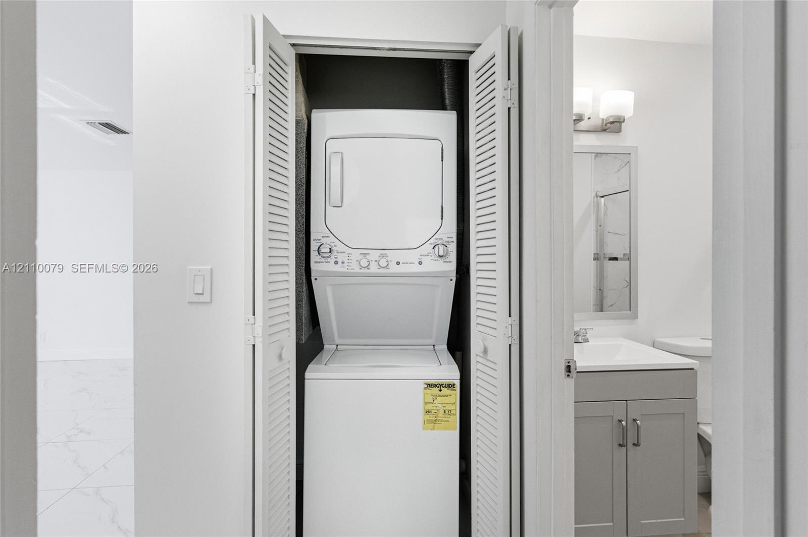 Laundry closet featuring louvered bi-fold doors, white stacked laundry unit, grey vanity cabinet with solid surface countertop, and wall-mounted vanity lighting
