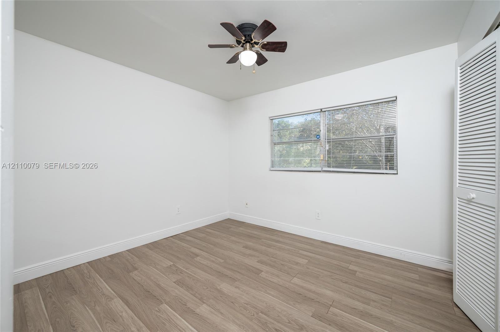 Neutral-toned wood-finish flooring, white walls, louvered bi-fold closet doors, and a ceiling fan with integrated lighting Neutral-toned wood-finish flooring, white walls, louvered bi-fold closet doors, and a ceiling fan with integrated lighting