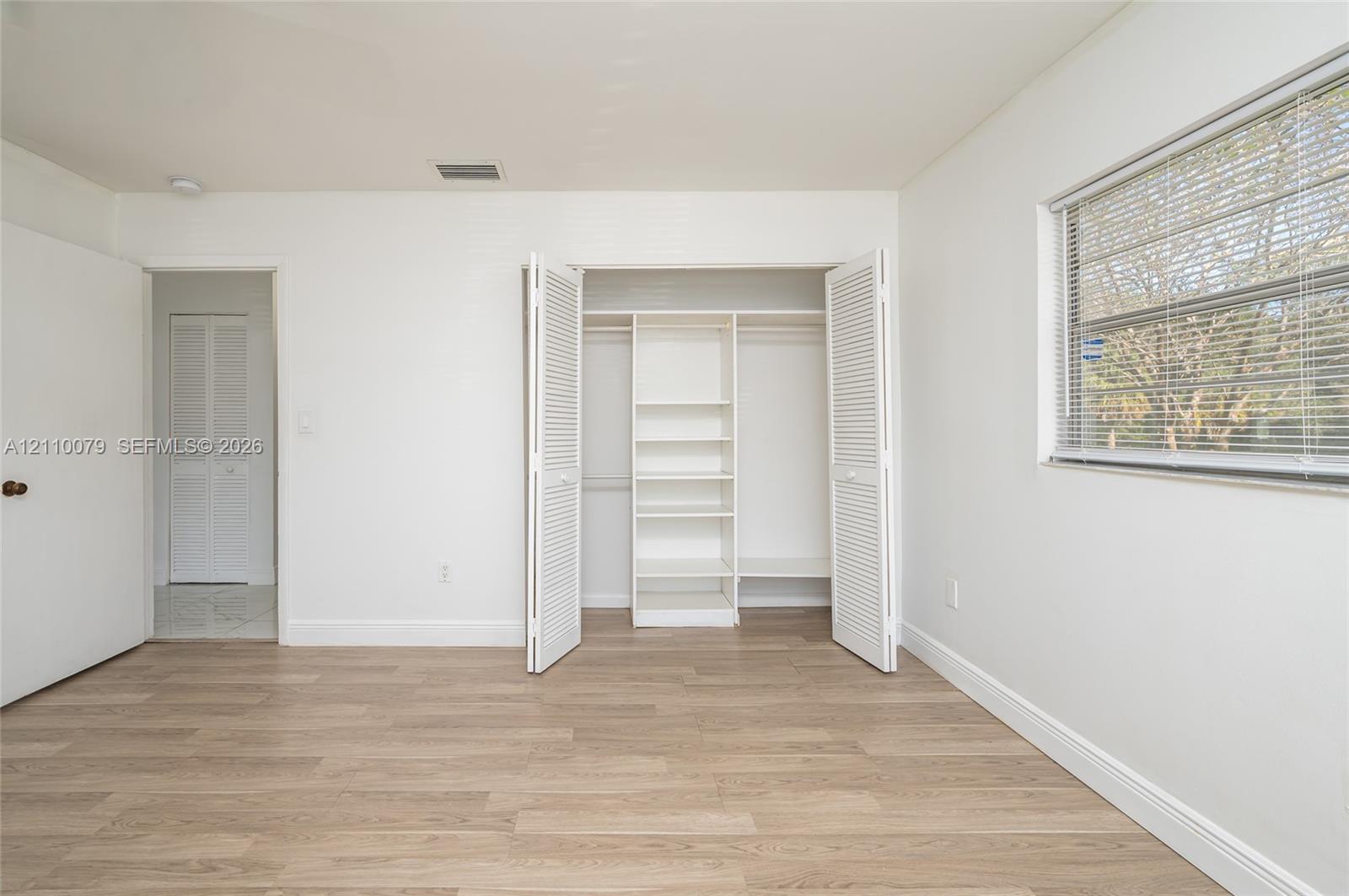 Room interior featuring wood-finish flooring, a window with horizontal blinds, and a closet with bifold doors and built-in shelving Room interior featuring wood-finish flooring, a window with horizontal blinds, and a closet with bifold doors and built-in shelving