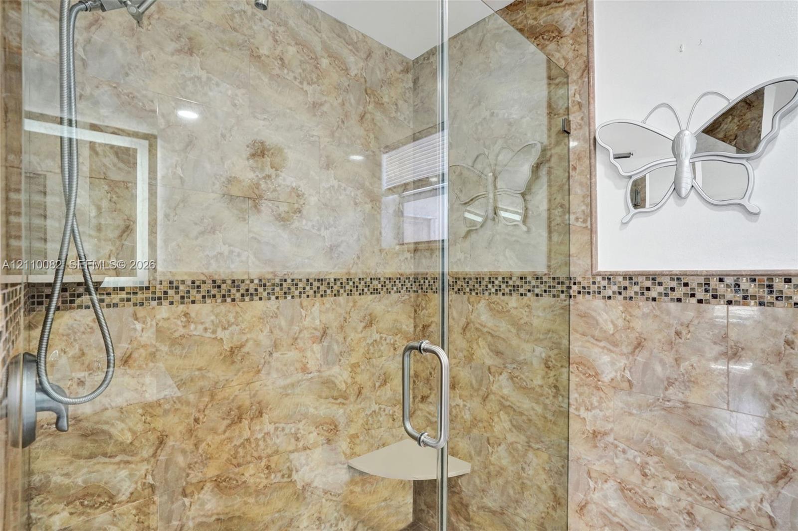 Walk-in shower featuring floor-to-ceiling stone-finish tile, glass enclosure, handheld shower fixture, built-in corner shelving, and decorative mosaic accent tile inlay
