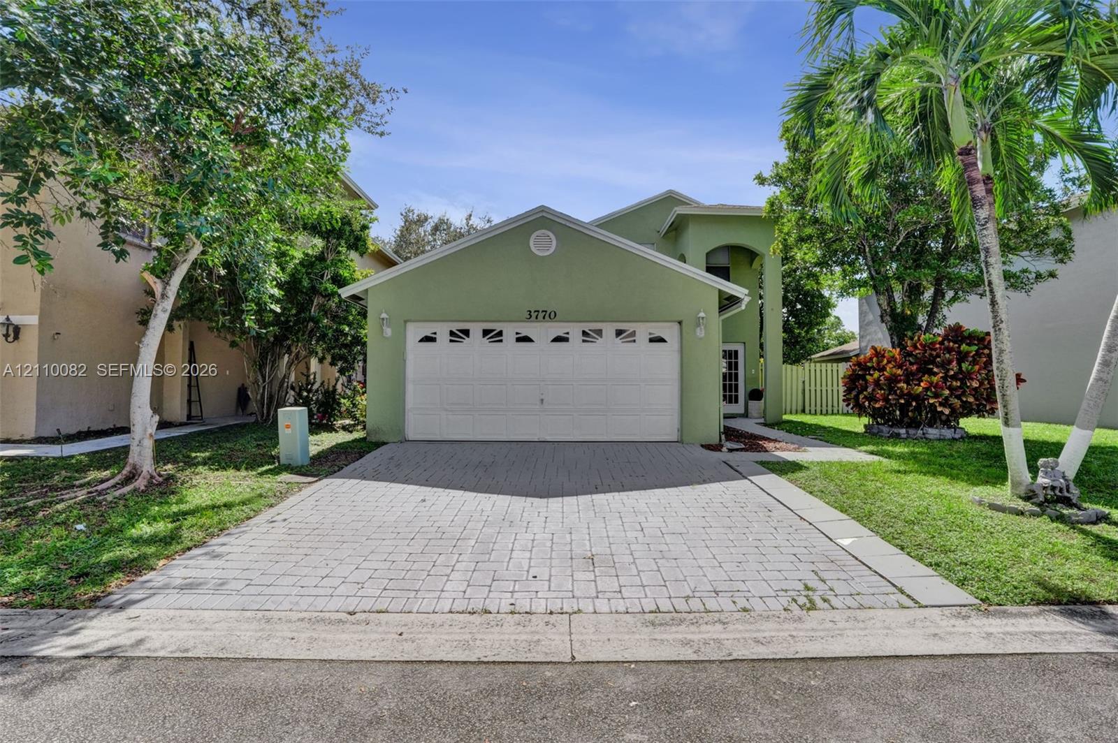Stucco exterior featuring a dual-car garage with arched decorative glass inserts and a paved stone driveway, surrounded by mature palm trees and landscaped shrubbery Stucco exterior featuring a dual-car garage with arched decorative glass inserts and a paved stone driveway, surrounded by mature palm trees and landscaped shrubbery