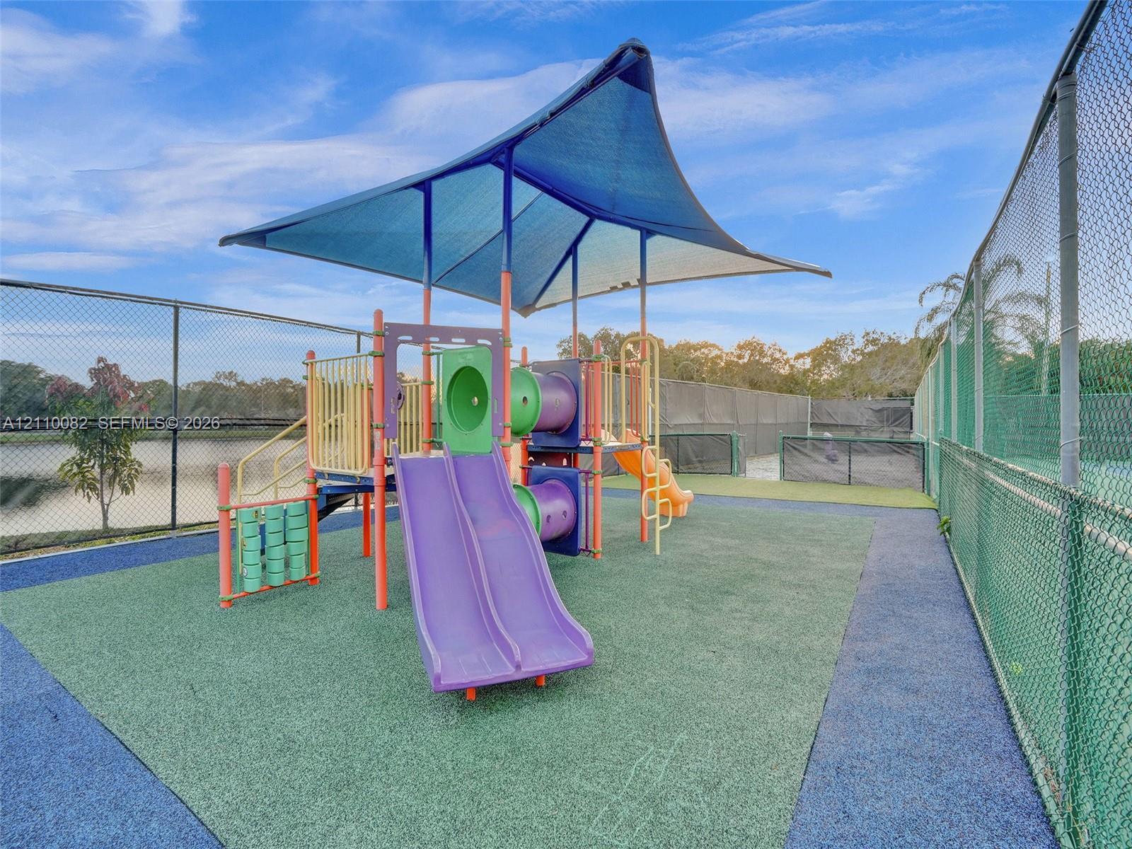 Recreational area featuring a playground structure with dual slides, a protective shade canopy, impact-absorbing surfacing, and perimeter chain-link fencing