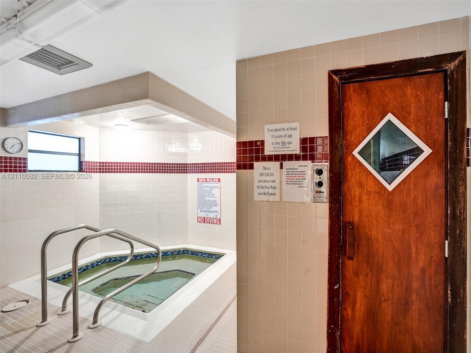 In-ground spa featuring metal handrails and white tiled surround, paired with a wooden sauna door equipped with a diamond-shaped viewing window
