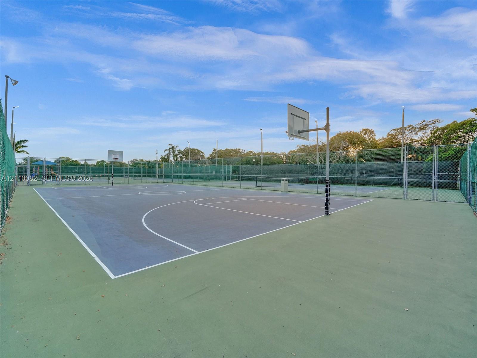 Outdoor sport court featuring a hard surface, dual basketball hoops, integrated perimeter fencing, and overhead light fixtures