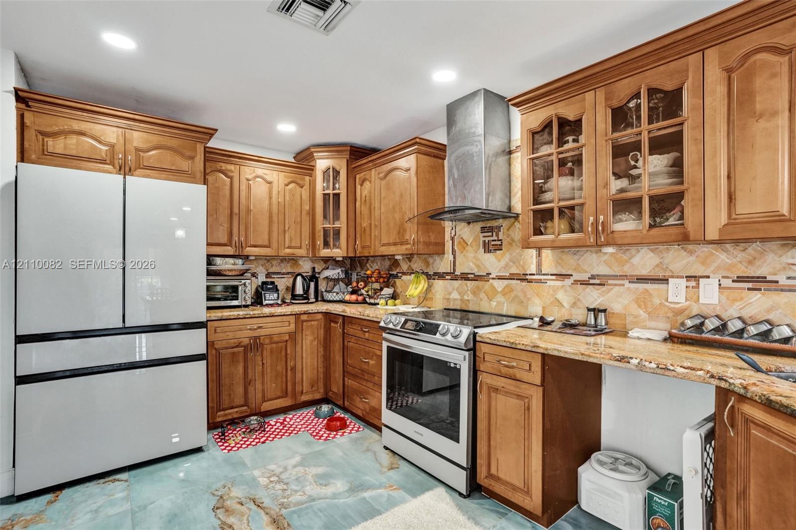 Kitchen featuring stained wood cabinetry with glass-front display accents, stone-finish countertops, herringbone-patterned tile backsplash, stainless steel range with a matching chimney-style hood, and large-format decorative floor tile