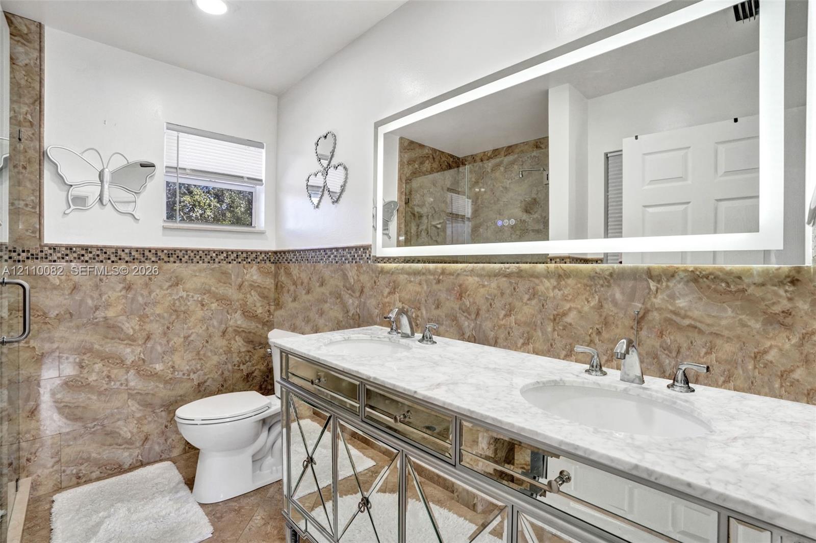 Double vanity with white stone countertop, dual undermount sinks, polished metal base, large LED-backlit mirror, and full-height stone tile wainscoting