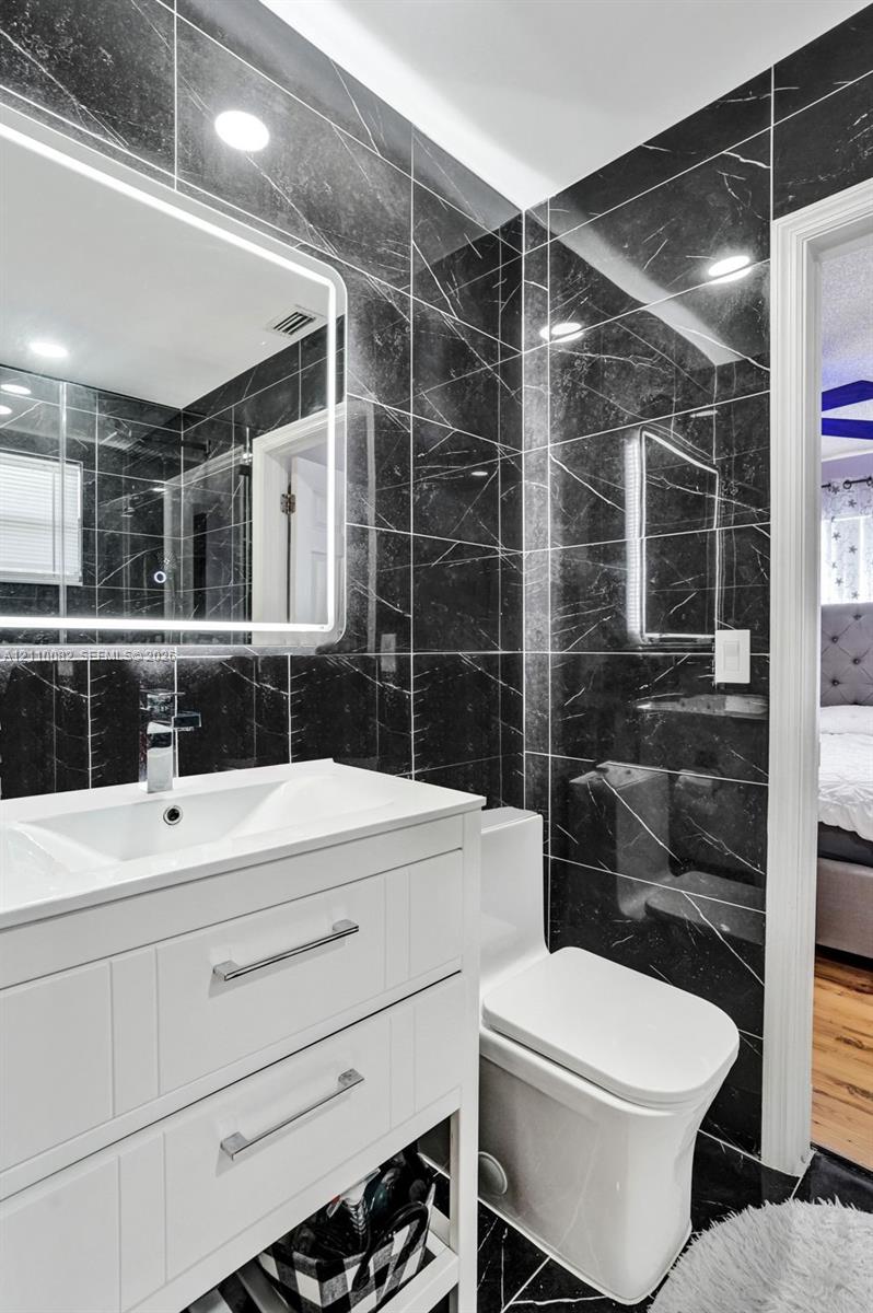 Bathroom featuring full-height dark stone-finish wall tile, white vanity with dual drawers, integrated rectangular sink, wall-mounted LED mirror, and modern elongated toilet